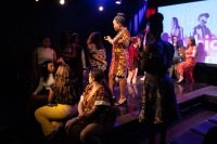 A woman stands on stage and speaks to models with a young model looking up at them