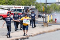 A man with a Energy Hi-Fives Here! sign holds their arm out to hi-five a firefighter with other firefighters in the background and a fire hydrant letting off water