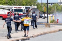 A man with a Energy Hi-Fives Here! sign holds their arm out to hi-five a firefighter with other firefighters in the background and a fire hydrant letting off water