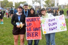 A pair of runners are seen with their friends who are smiling at the camera holding up their signs which say "Hurry up, we want beer" and "Run Dunn run!"