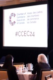 Professionals sit at a table in front of a projection showing the Chamber of commerce Executives of Canada logo and #CCEC24