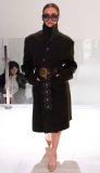A model walks on the runway wearing a coat with a large round metal ornament on their belt