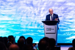 A man at the podium gestures to the crowd seen in the foreground