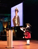 A man in historic cap and gown is seen reading from a scroll with their image projected on a large screen