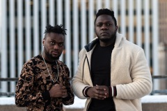 Two people look into the camera with their hands together in front of a blurred building facade