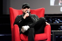 A man smiles while sitting on a red chair on stage