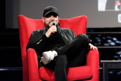A man smiles while sitting on a red chair on stage