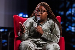 A person sits in a red chair and gestures while speaking
