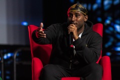 A man gestures while speaking and sits on a red chair