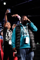 A teenager smiles on stage while rapping into the microphone with two teenagers taking a selfie behind him