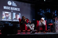 A man speaks on stage with another listening to them and Music Business is displayed behind them